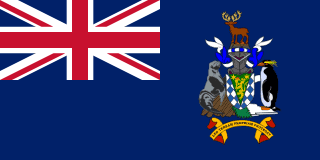 The flag of South Georgia is blue with the UK flag in the canton. The national coat of arms is centered on the fly half and has a green shield with a golden lion holding a torch. A fur seal is to the left of the shield and a Macaroni penguin to the right. A reindeer appears above the crest, and below the shield on a scroll is the Latin motto "LEO TERRAM PROPRIAM PROTEGAT" (Let the Lion Protect its Own Land).